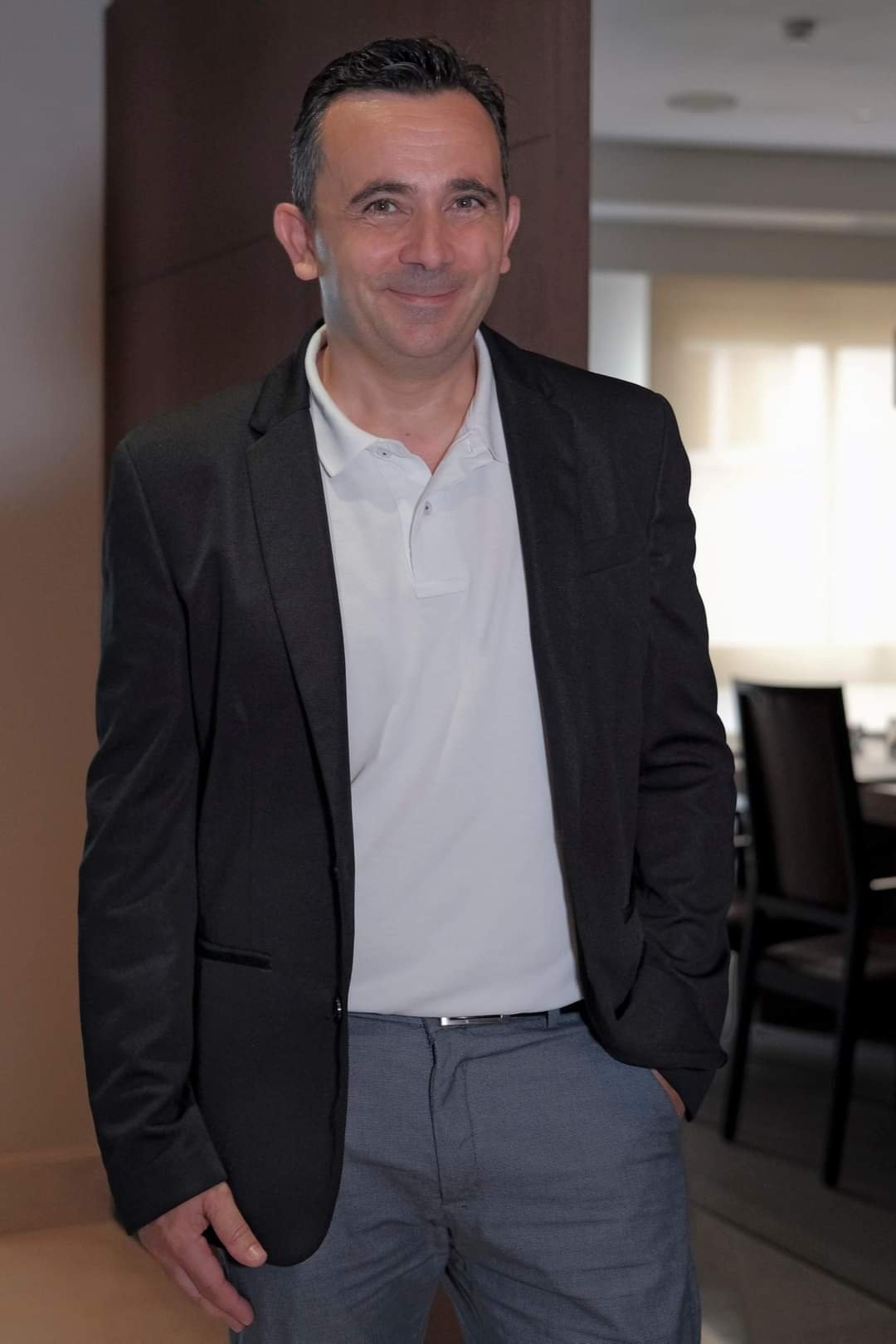 Hombre con saco oscuro, camiseta tipo polo blanca y pantalones grises posando y sonriendo en un interior iluminado con mesas y sillas al fondo.
