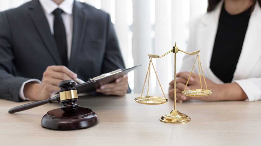 Mazo de juez y balanza dorada sobre un escritorio, con un hombre y una mujer vestidos formalmente sentados al fondo; el hombre sostiene una carpeta.