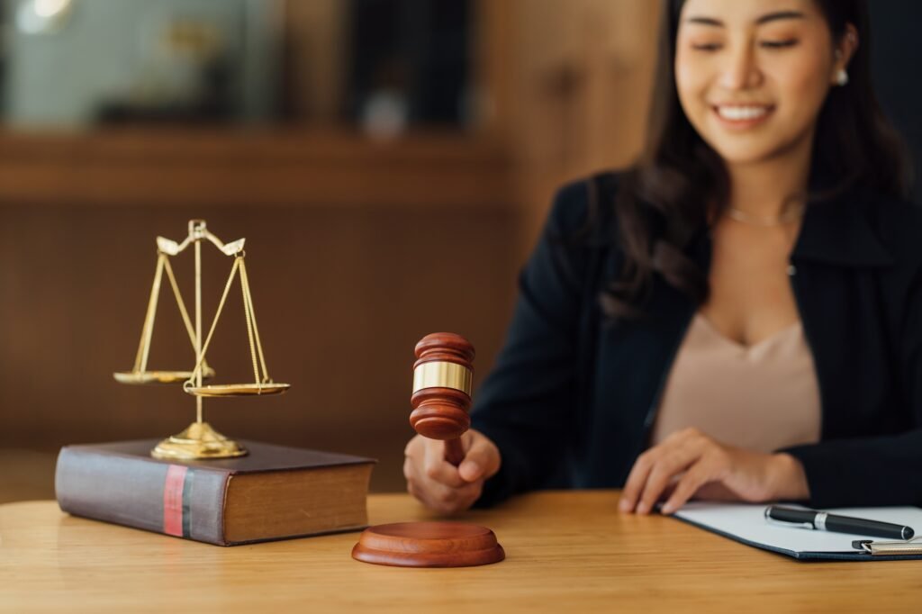 Mujer sonriendo mientras sostiene un mazo de juez sobre una mesa; junto a ella hay una balanza dorada sobre un libro grande y una carpeta con bolígrafo.